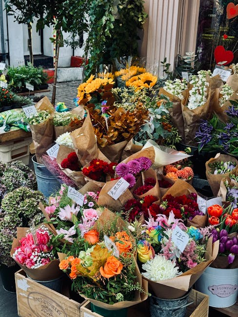 A person wearing a white t-shirt with visible tattoos on their arm, seen from the back, is carrying a large bouquet of fresh flowers wrapped in brown paper towards a flower delivery shop. The bouquet