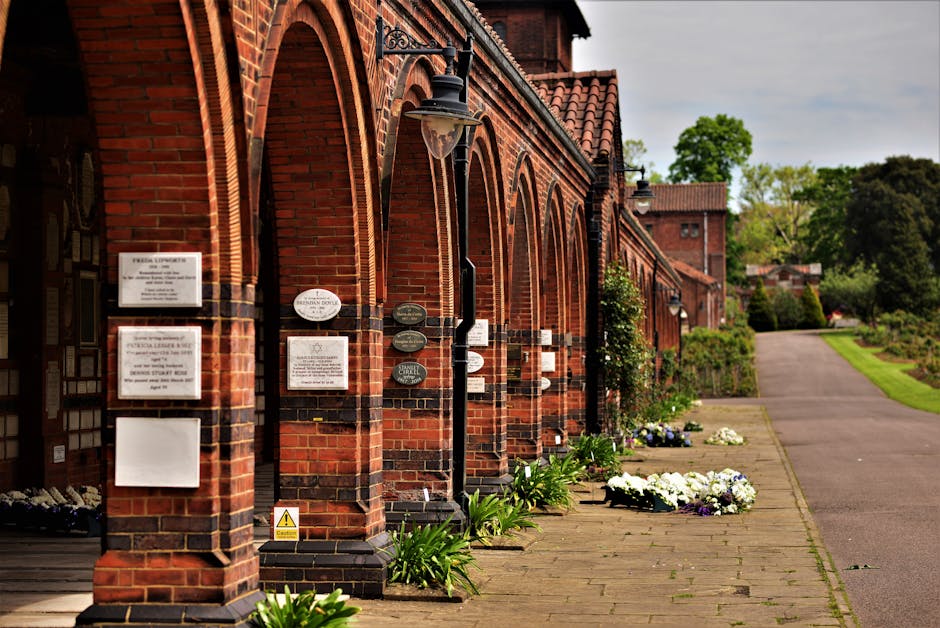 A group of three people, two women and one man, walking through a cemetery on a sunny day. The women are holding a floral arrangement with white and green flowers, wrapped in transparent and white pap