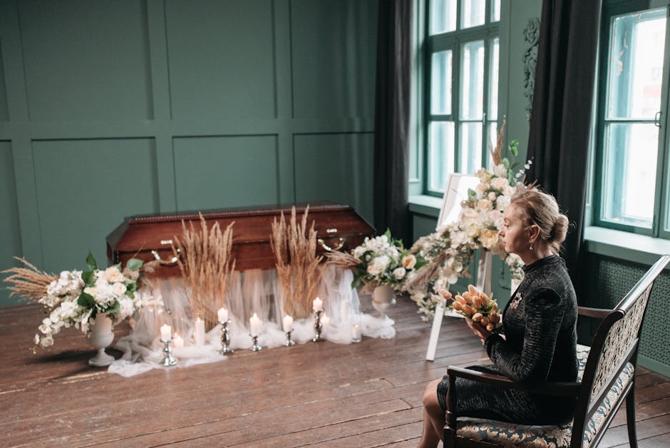 A large, cascading floral arrangement resting atop a wooden casket, featuring a mixture of roses, lilies, and other flowers with varied petal textures. The bouquet includes both open and tightly close